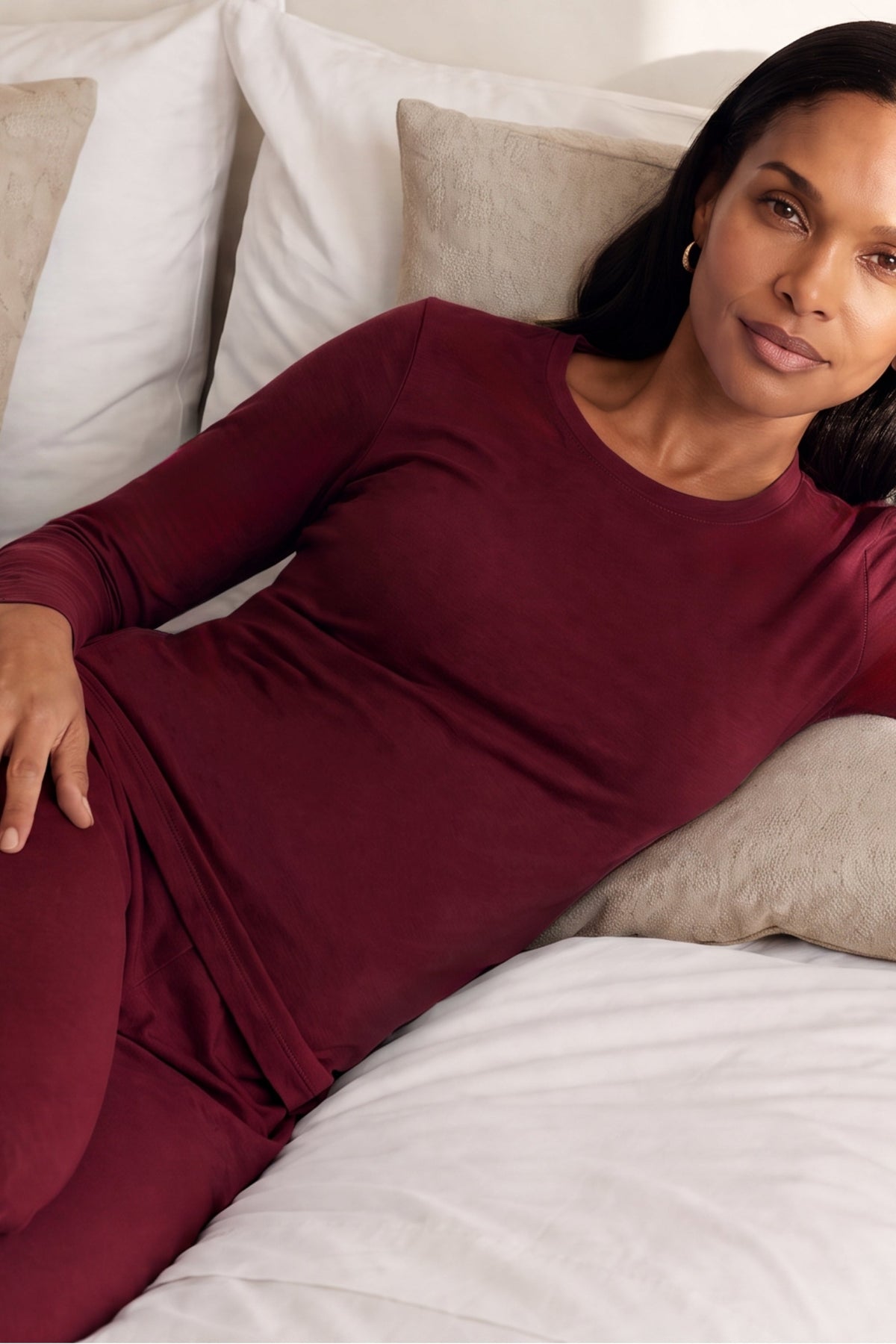 A woman reclining on a bed wearing a matching burgundy long-sleeve pajama set against white and beige pillows.