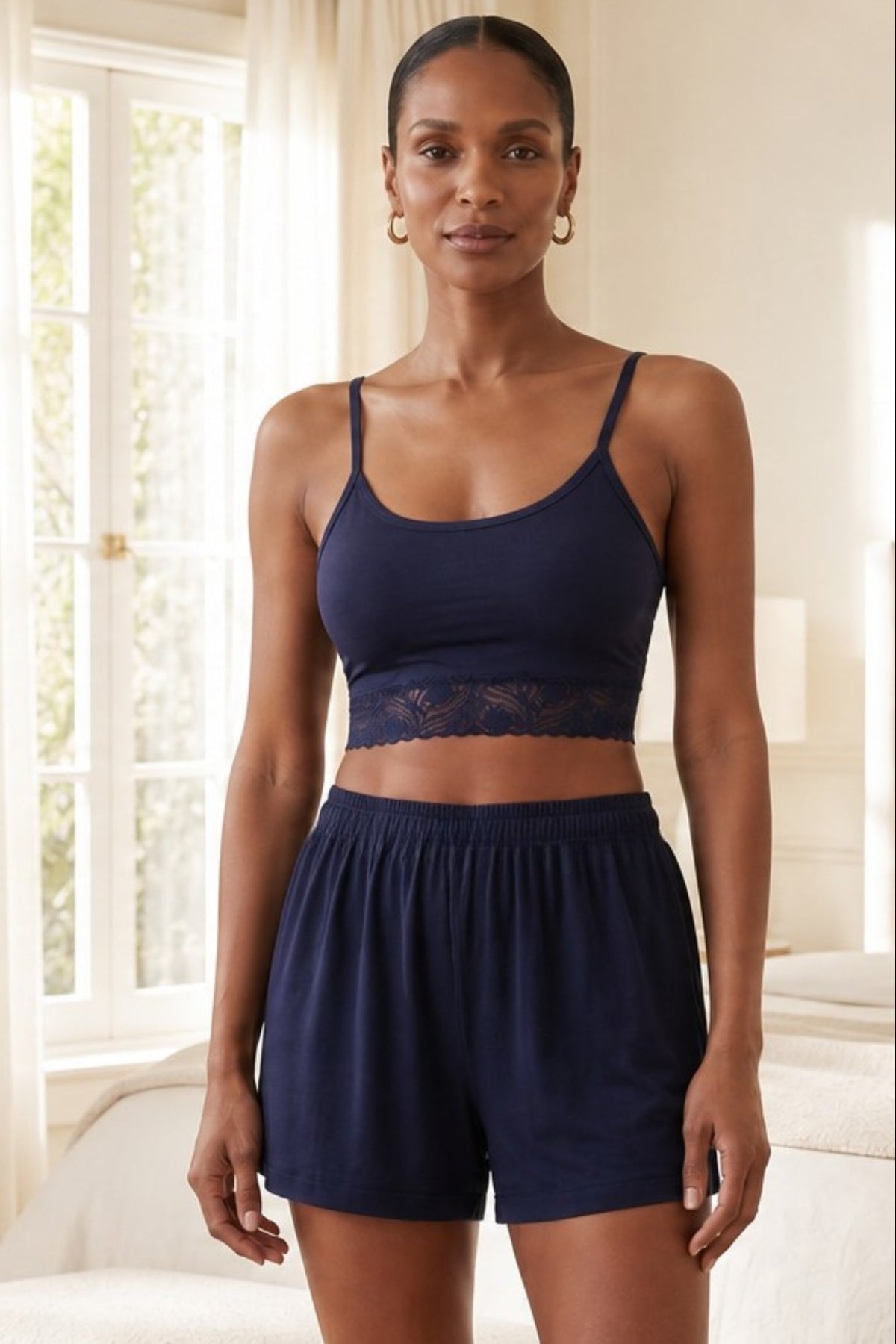 A woman modeling a matching navy blue loungewear set consisting of a lace crop with a lace band and matching bamboo shorts, standing in a brightly lit bedroom.