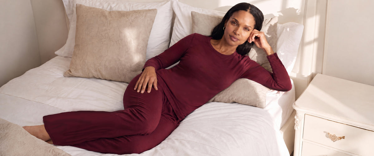 A woman reclining on a bed wearing a matching bordeaux long-sleeve pajama set with wide-leg pants, surrounded by white bedding and beige pillows.
