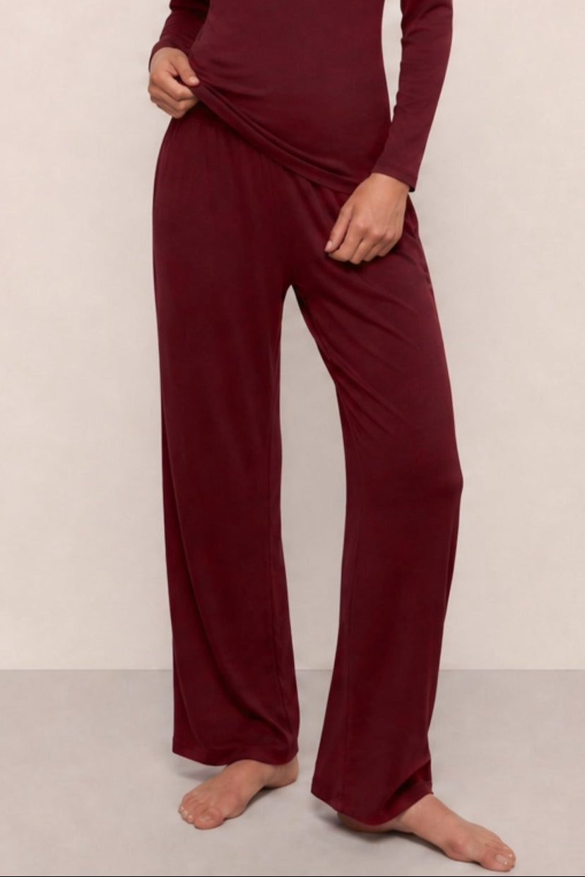 A woman modeling a long-sleeve, crew-neck and lounge pants in a deep burgundy color, standing against a neutral beige studio backdrop.
