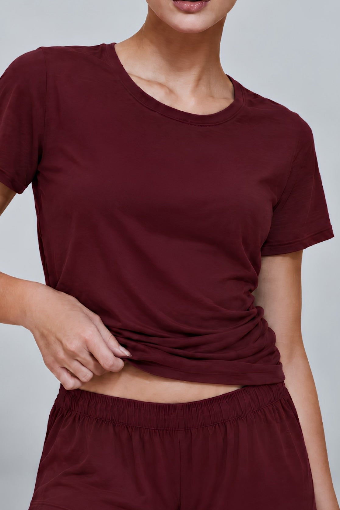A woman wearing a matching burgundy short-sleeve crewneck t-shirt and shorts set, shown from the chin down against a plain light gray studio background.
