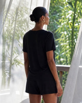 Person wearing camofleur relaxed tee and shorts in noir black standing in a room with sheer curtains and bamboo outside