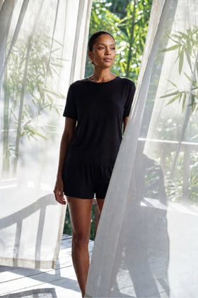 Woman in a black bamboo lounge set from camofleur standing in front of sheer curtains with greenery outside